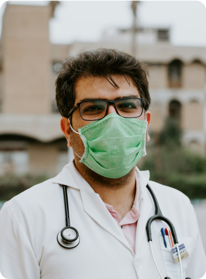 Young male physician looking at the camera with a muted green face mask and stethoscope around his neck. 